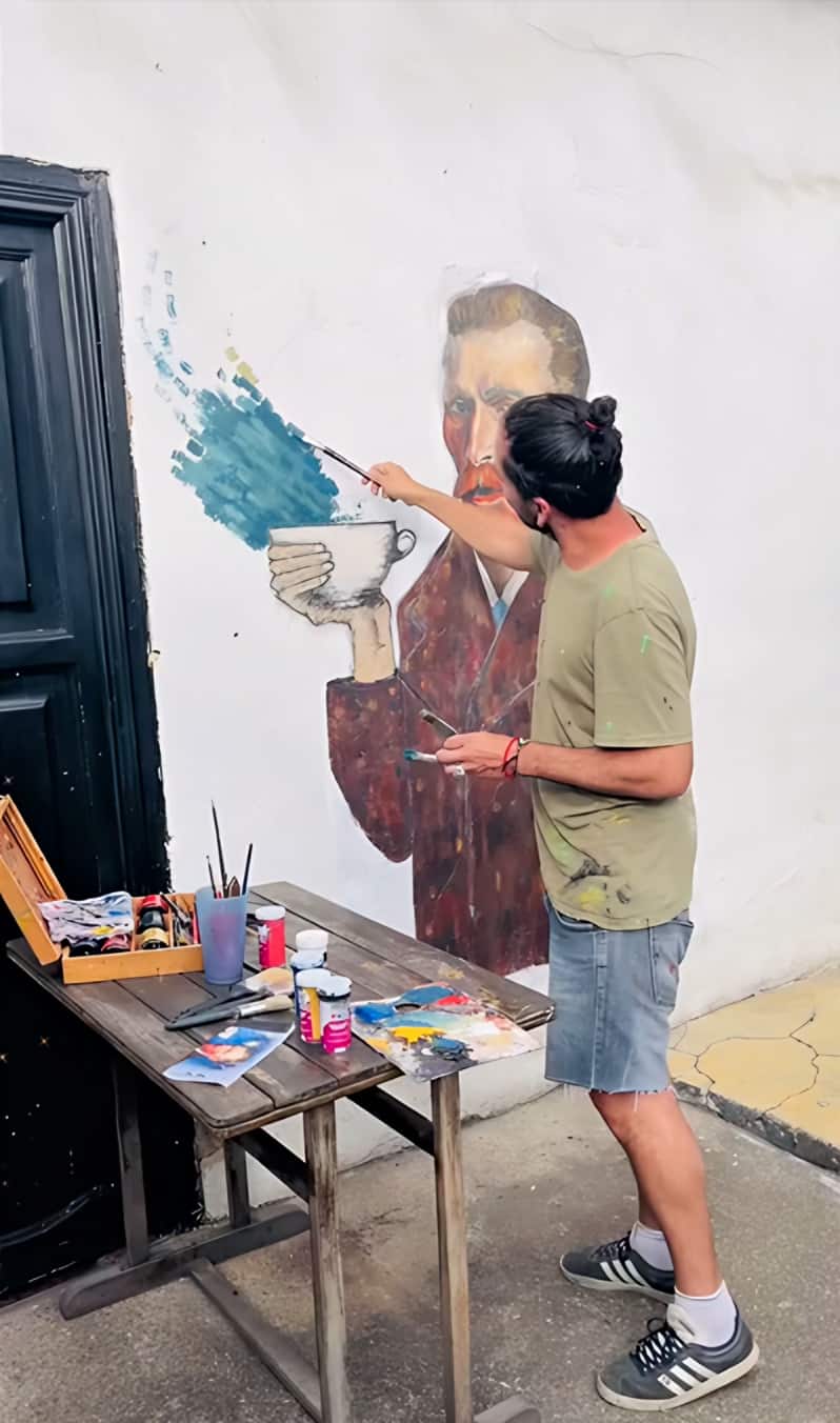 Artista pinta el mural de Van Gogh con taza junto a una puerta negra, con su mesa de pinturas al lado
