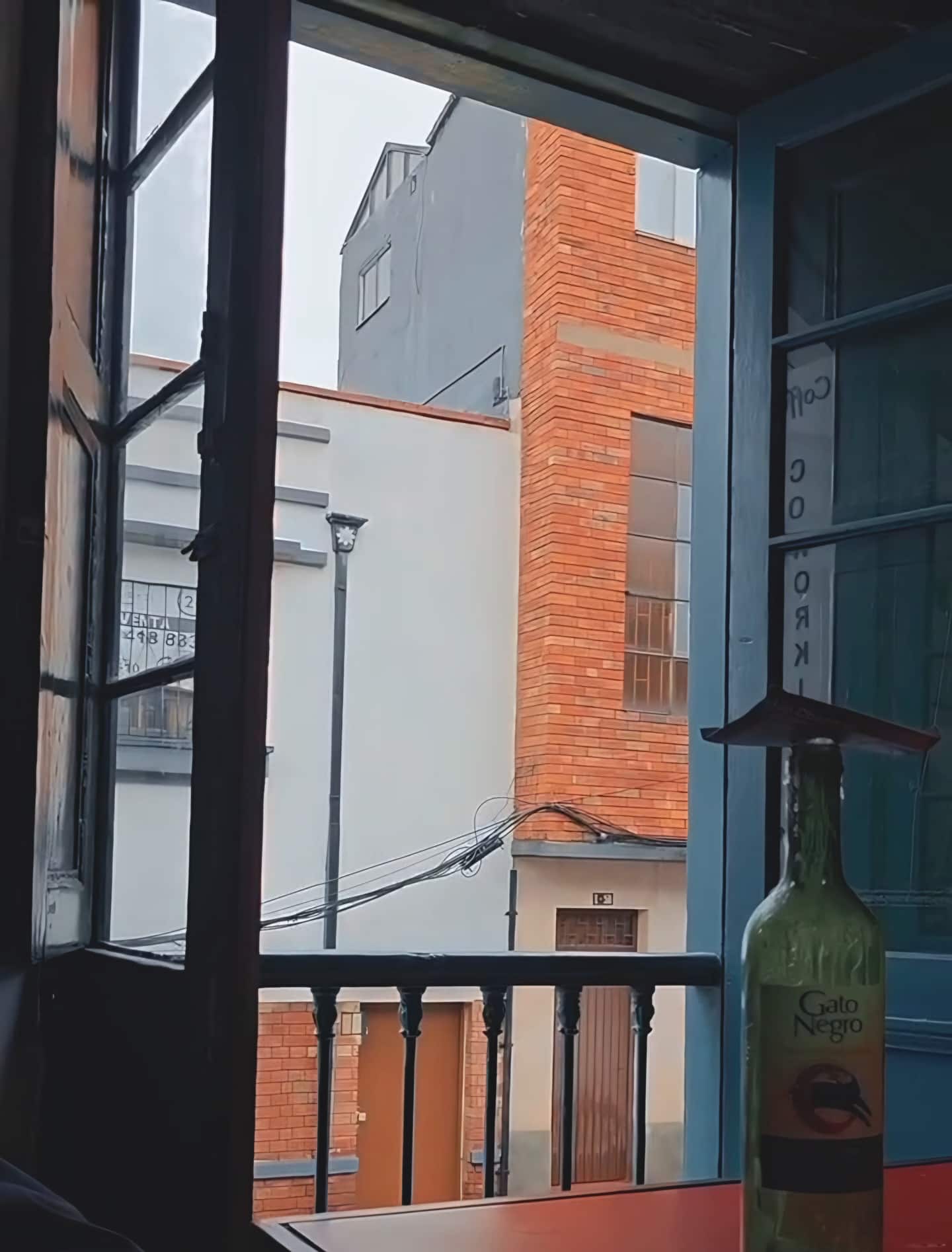 Ventana abierta con baranda hacia la calle y casas de ladrillo, junto a una botella sobre una mesa roja