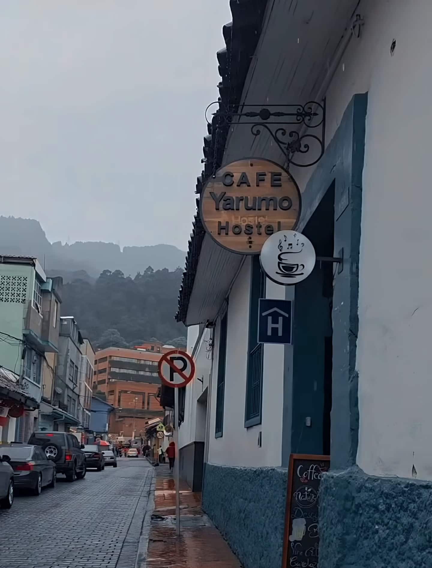 Calle 12 mojada por la lluvia, con el aviso «Café Yarumo Hostel» en la fachada y cerros entre la niebla