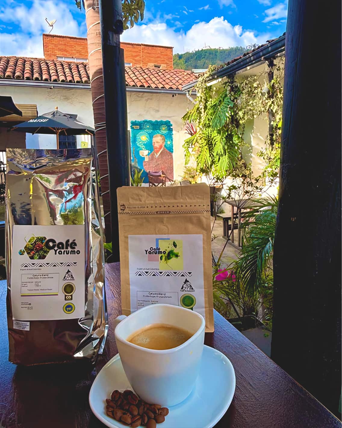 Dos bolsas de Café Yarumo Caturra Blend y una taza de café en el patio, con el mural y los cerros al fondo