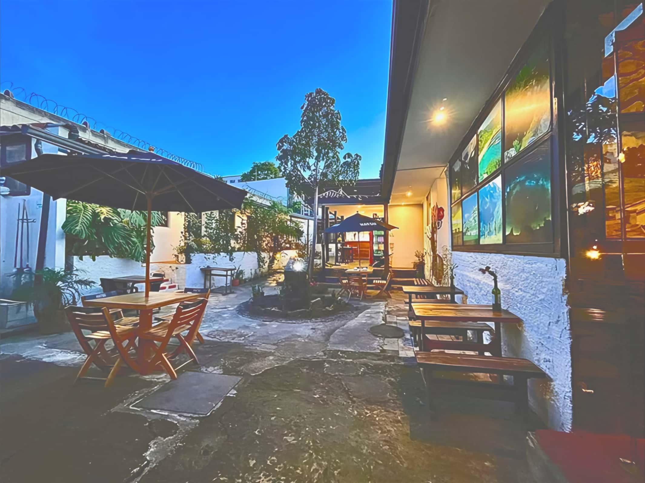 Patio colonial al anochecer con sombrillas, mesas de madera, bancas y una fuente con árbol bajo un cielo azul