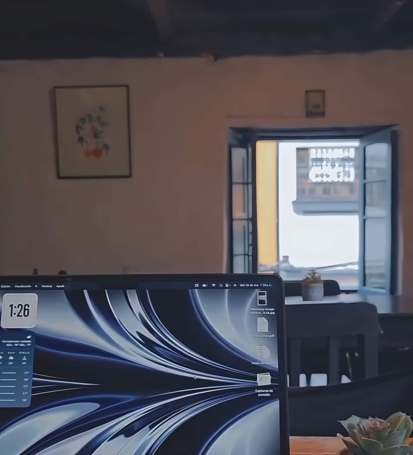 Portátil abierto en una mesa de madera frente a una ventana del salón, con un cuadro en la pared