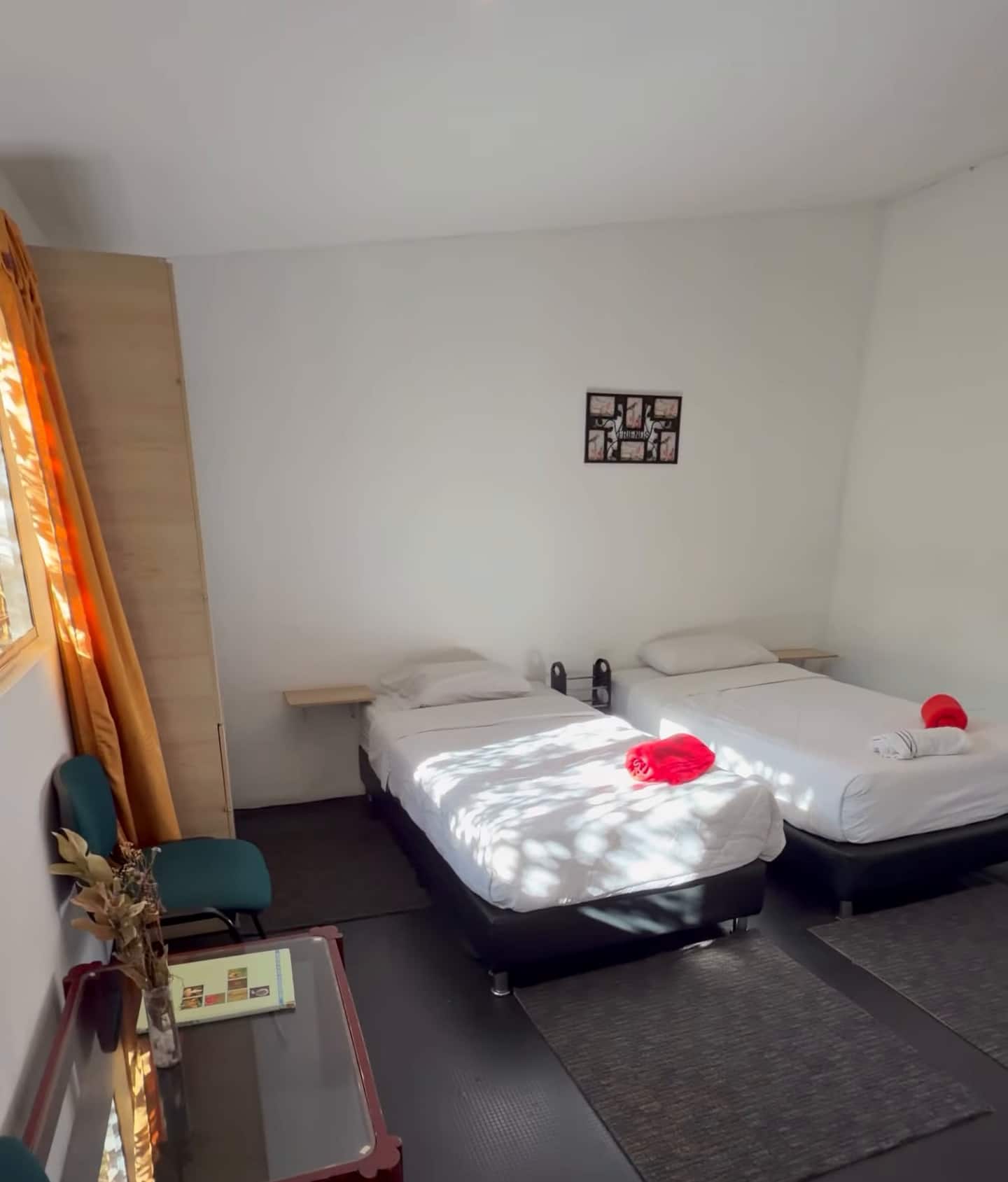 Habitación luminosa con dos camas sencillas, piso oscuro y un tapiz rojo en la pared