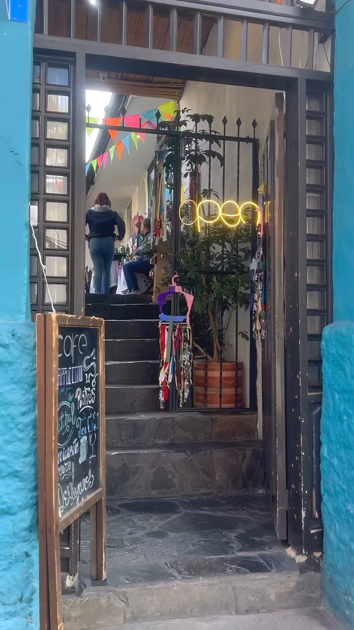 Entrada turquesa con reja abierta, escaleras de piedra, tablero de tiza y letrero luminoso «open»