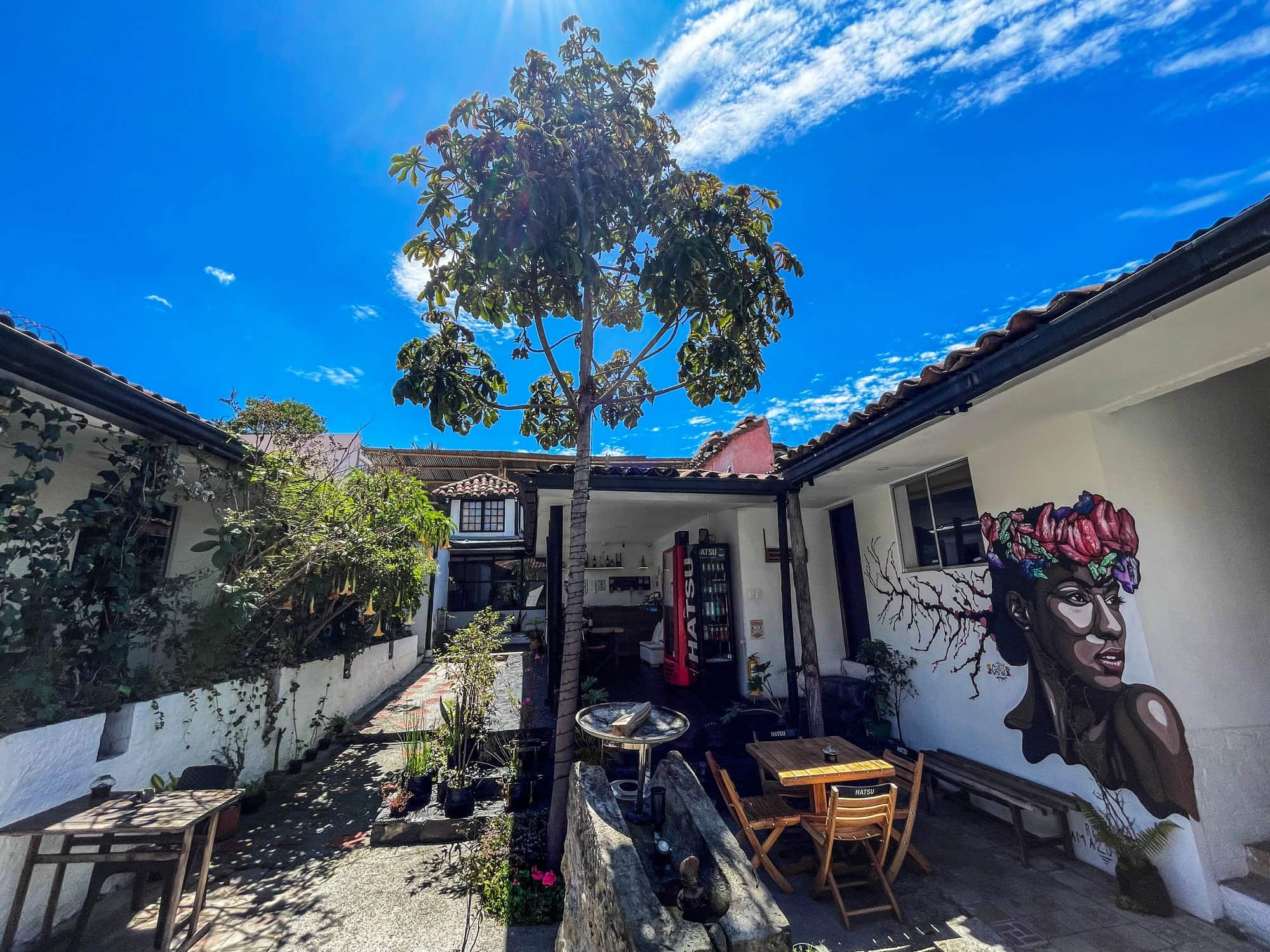 Patio con árbol alto, mesas y mural de mujer con flores