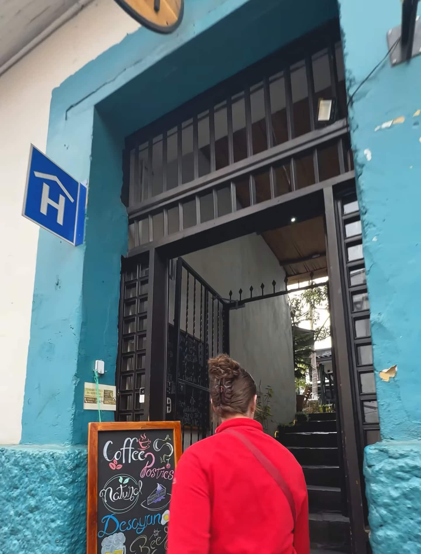 Una visitante entra por la puerta turquesa del hostal, con reja negra, escaleras y tablero de café