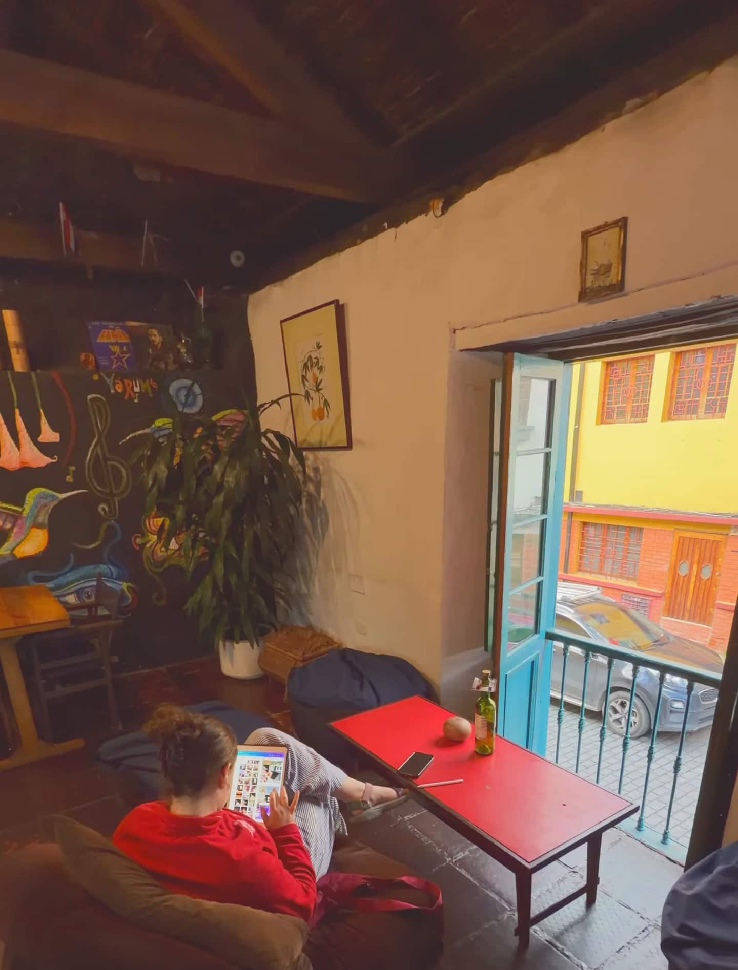 Salón con techo de madera y ventana abierta a la calle; una huésped lee una tableta junto a una mesa roja