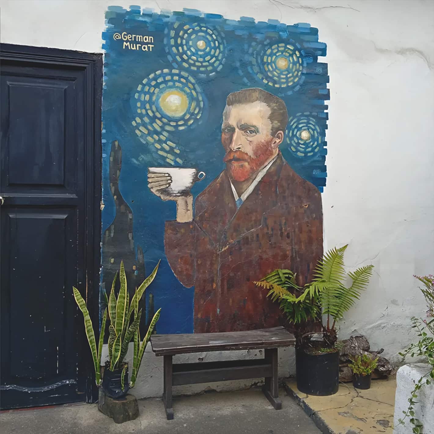 Mural de Van Gogh sosteniendo una taza de café, firmado @German Murat, con una banca de madera y plantas