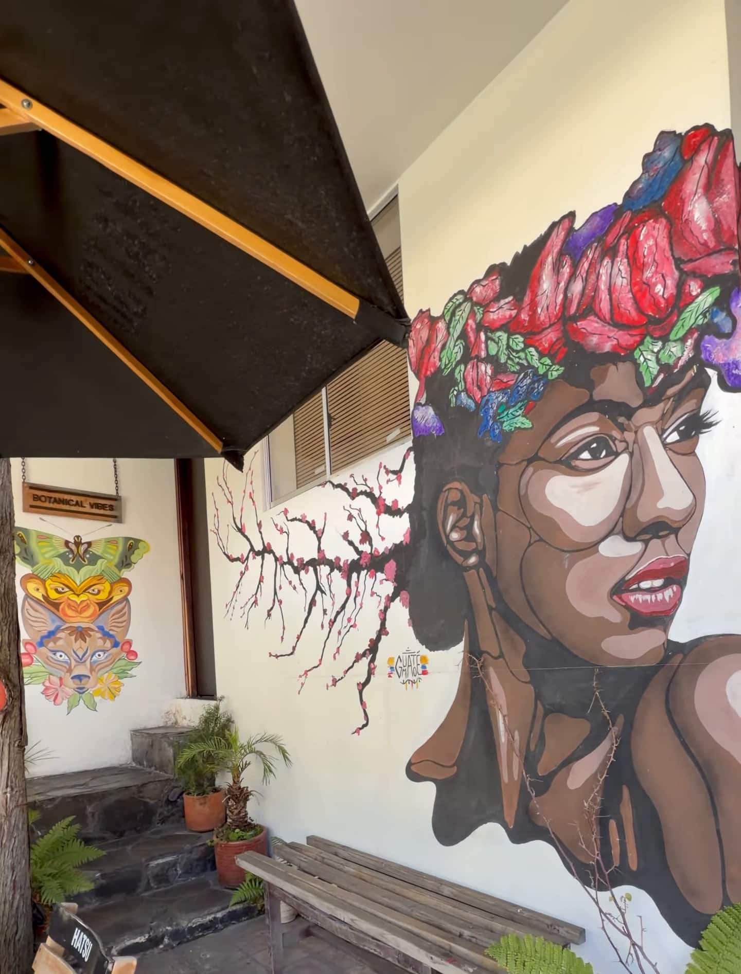 Mural de una mujer con corona de flores en un muro del patio, sobre una banca de madera