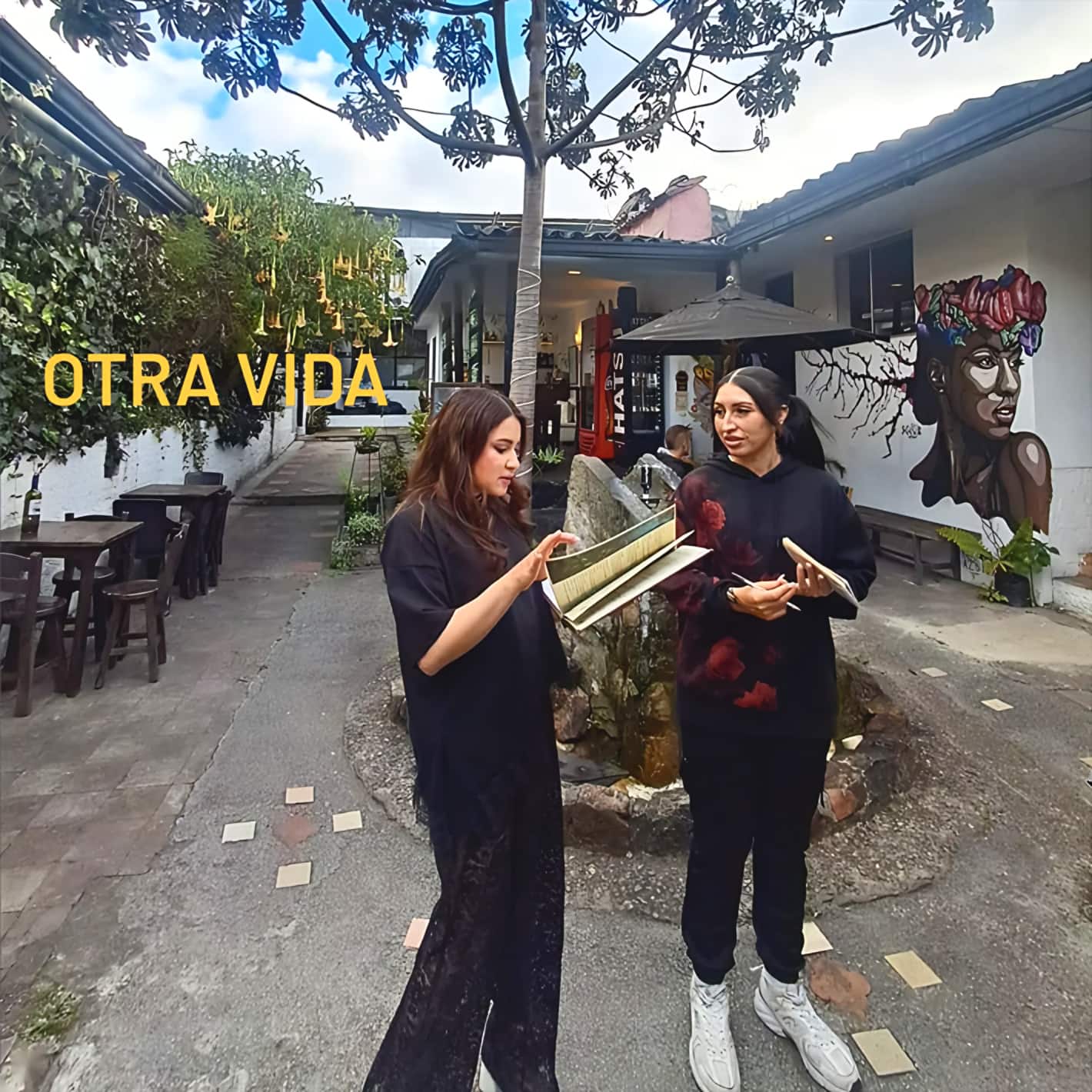 Dos mujeres revisan una partitura en el patio, junto a la fuente y un mural; texto amarillo «OTRA VIDA»