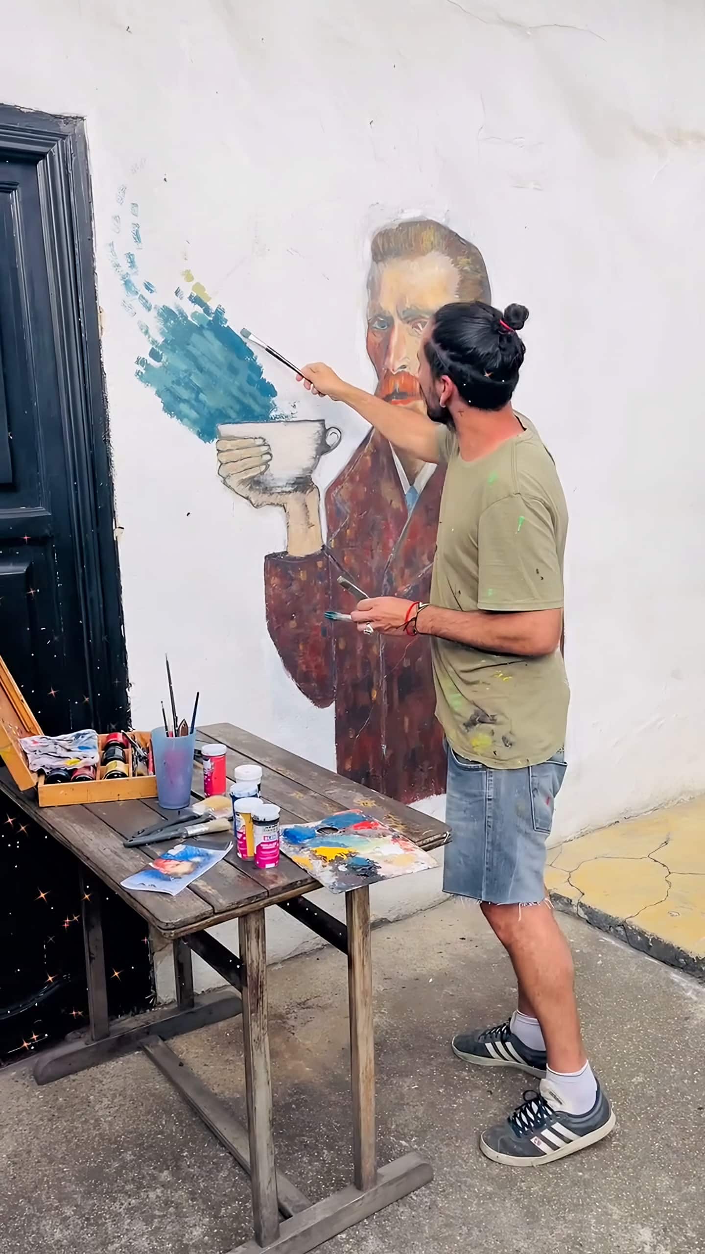Artista pinta los trazos azules del mural de Van Gogh, con su mesa de pinturas junto a la puerta