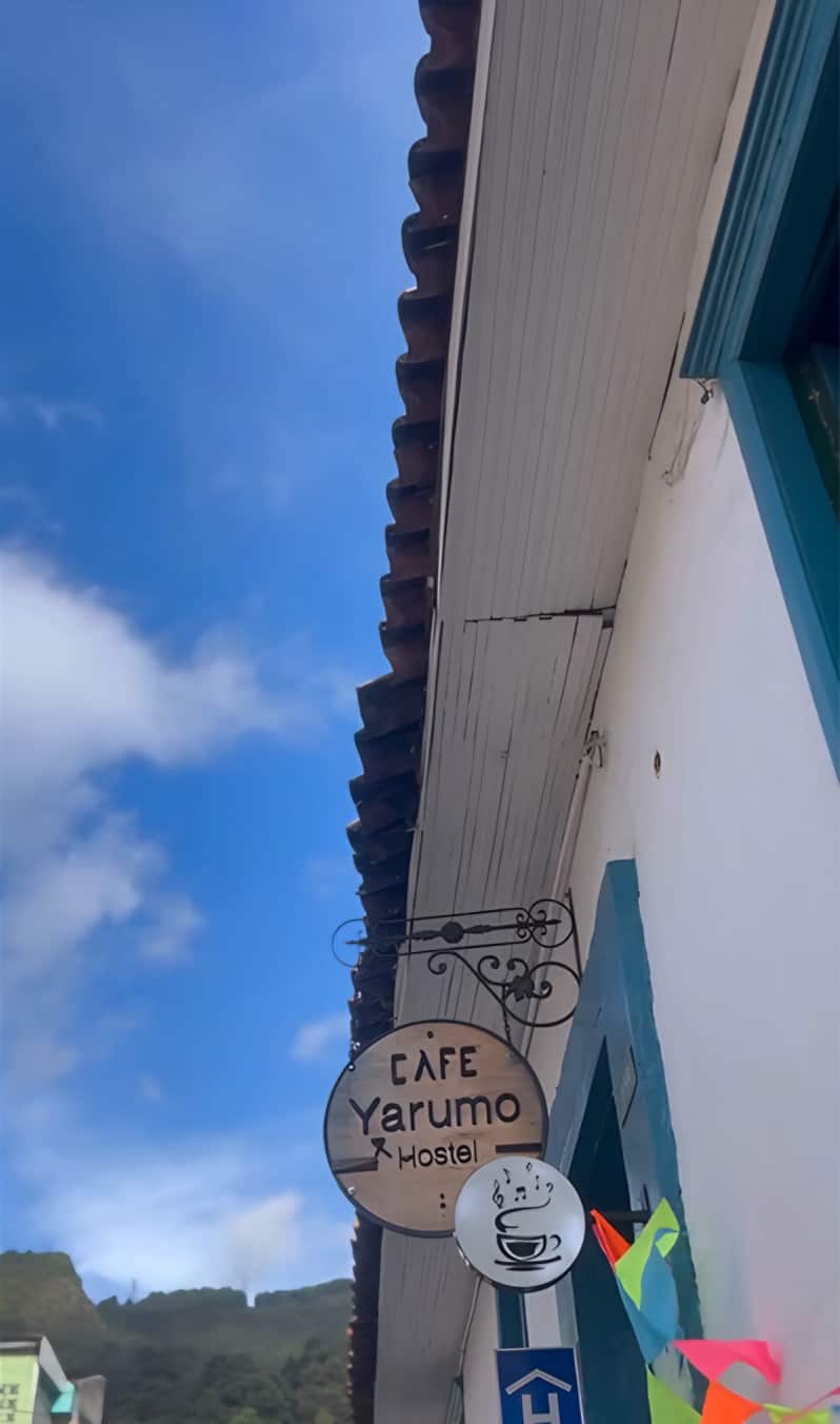Aviso de hierro «Café Yarumo Hostel» colgado bajo el alero de teja, con cielo azul y nubes