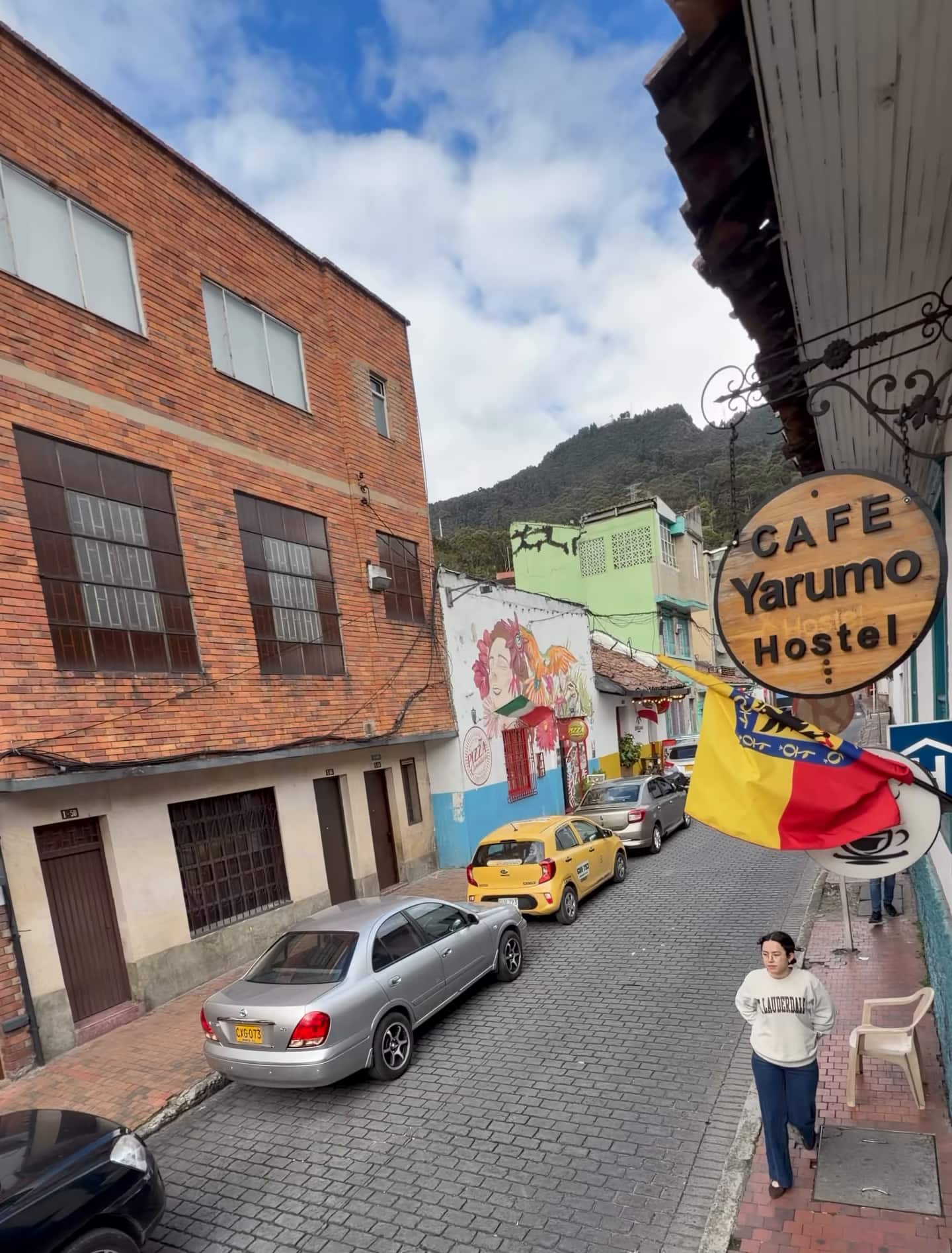 Aviso «Café Yarumo Hostel» y bandera sobre la calle empedrada, con casas de colores y los cerros detrás