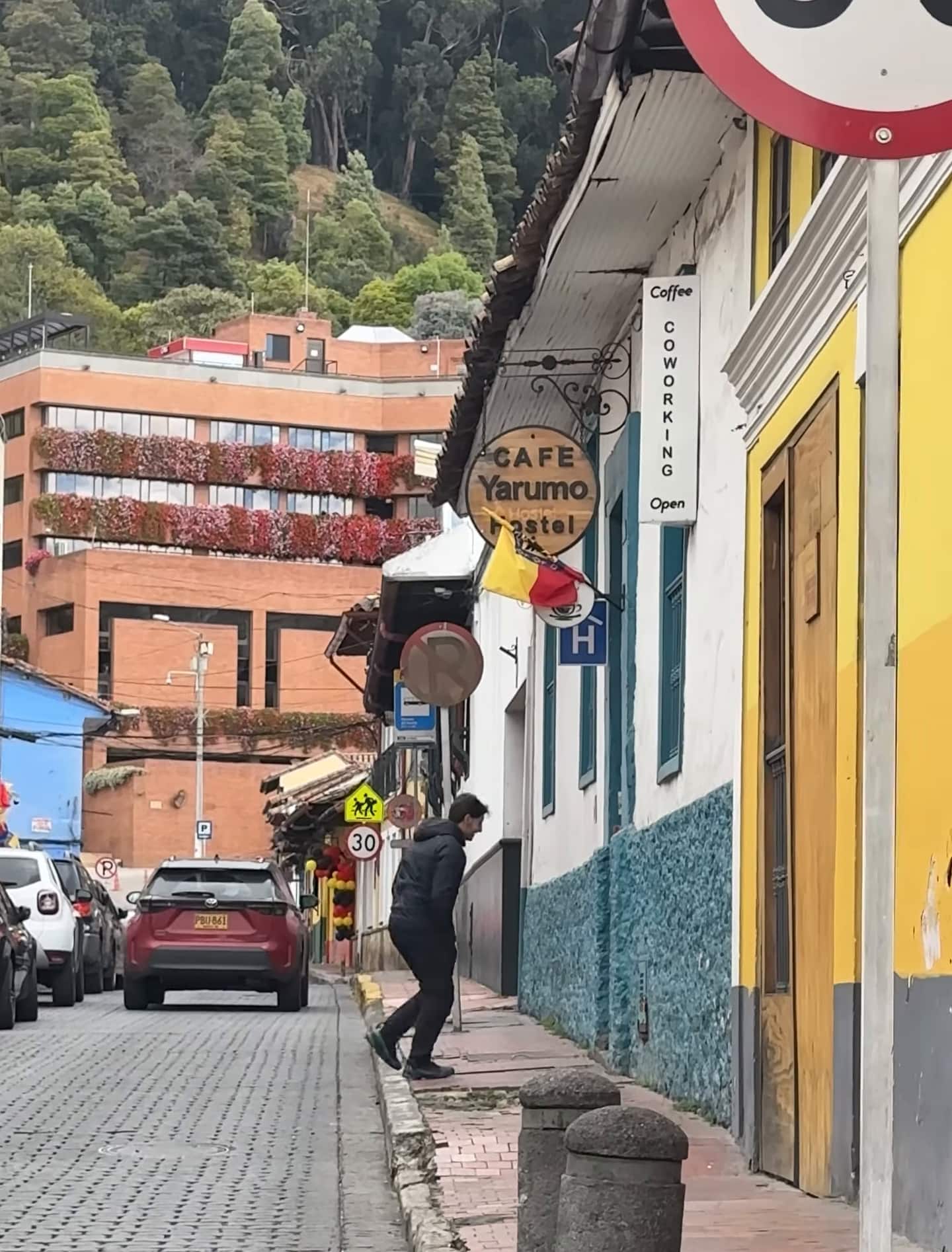 Calle empedrada de La Candelaria con el aviso «Café Yarumo» en una fachada y los cerros arborizados al fondo