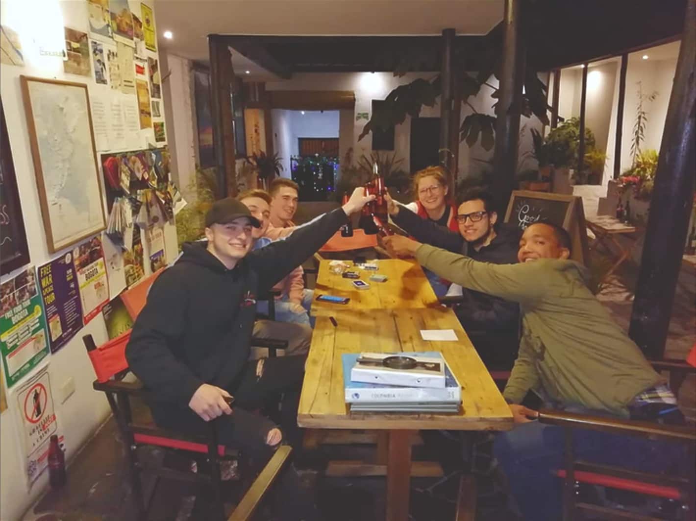 Huéspedes brindan con cervezas en una mesa de madera del salón común, con afiches de viaje en la pared
