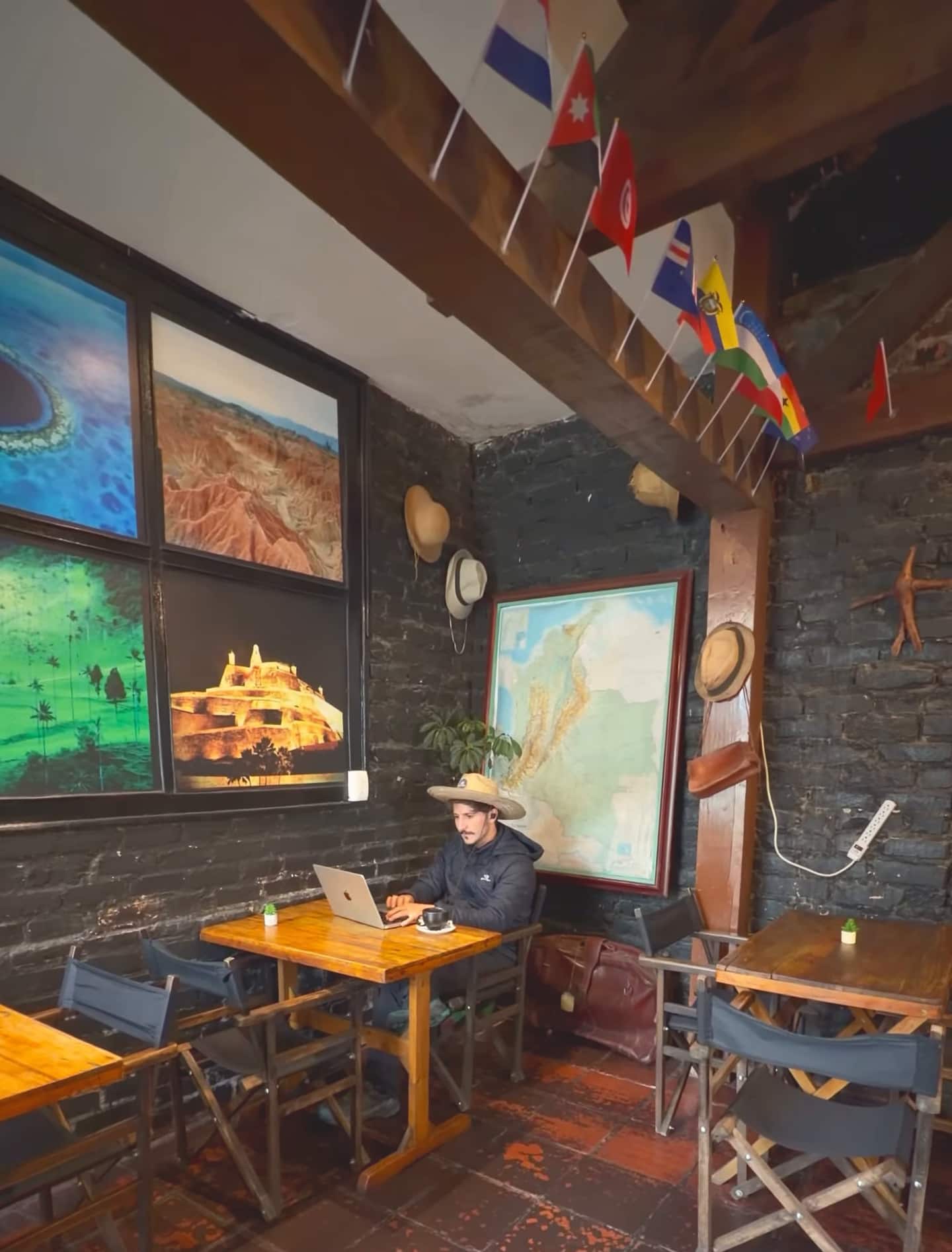 Hombre con sombrero trabaja en su portátil en un salón de muro de piedra, con mapa, afiches y banderines